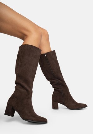 Botas de ante marrón hasta la rodilla con tacón de bloque, que cuentan con un cierre lateral, puntera afilada y un diseño ligeramente arrugado.
