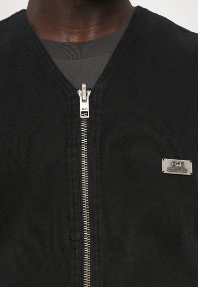 Jachetă din denim negru, cu închidere cu fermoar argintie și un mic etichetă metalică cu logo. Material texturat, cu un design casual și ajustat, având decolteu în formă de V.