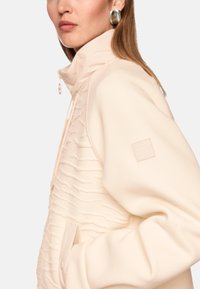 Cream-colored jacket with textured, wavy fabric on the front, a high collar, and a small logo patch on the left shoulder. Smooth finish.
