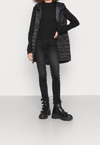 Un gilet nero imbottito con cappuccio sopra un maglione aderente nero a coste, abbinato a jeans skinny neri e stivali da combattimento neri chunky con cinturini.