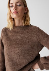 Brauner flauschiger Pullover mit hohem Kragen und lockerem Sitz. Die Textur ist weich mit einer leicht zerzausten Optik und breiten Ärmeln.