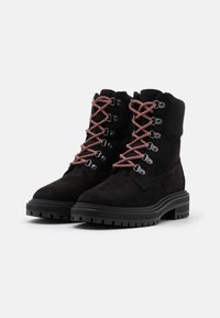 Botas de tobillo de ante negro con suelas de goma gruesas y cordones de patrón rojo y blanco pasados por ojales de metal.