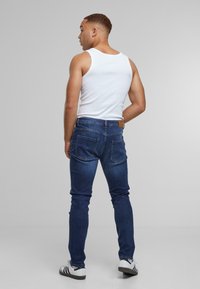 Un débardeur blanc sans manches et un jean bleu foncé moulant sur une personne, avec une texture lisse et un design standard à cinq poches, vu de derrière.