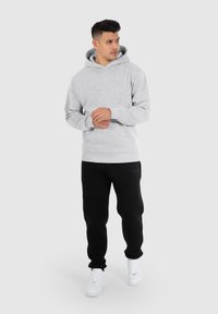 Grauer Hoodie mit Kängurutasche, getragen mit schwarzen Jogginghosen und weißen Turnschuhen. Der Hoodie hat eine Kordel und gerippte Bündchen.