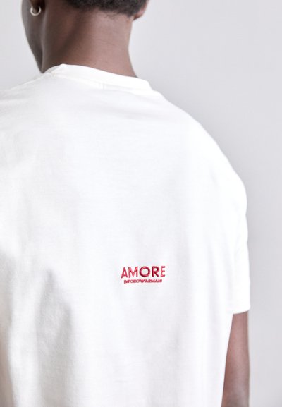 Valge puuvillane T-särk, millel on punane tikandiga tekst "AMORE" ja "EMPORIO ARMANI" seljaosas. Ümar kaelus ja lühikesed varrukad. Pehme tekstuur.