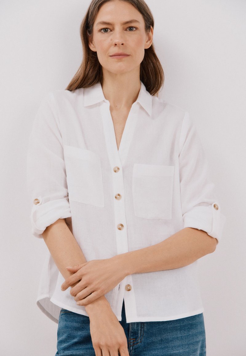 Camicia bianca in lino con due tasche frontali, bottoni frontali in una tonalità chiara, e maniche arrotolate fissate con bottoni in tono oro.