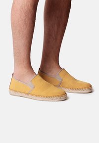 Scarpe gialle senza lacci realizzate in tessuto strutturato con accenti elastici color beige, dotate di una suola intrecciata in juta per aderenza e stile.