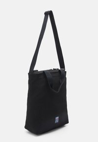 Zwarte draagtas met een gladde canvasstructuur, voorzien van geweven handvatten, een ritsafsluiting en een klein merkembleem aan de voorkant.