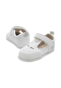 Zapatos de bebé blancos con puntera redondeada, diseño de tira en T, parte superior perforada y suela suave y texturizada. Cuenta con una correa de Velcro para un ajuste seguro.
