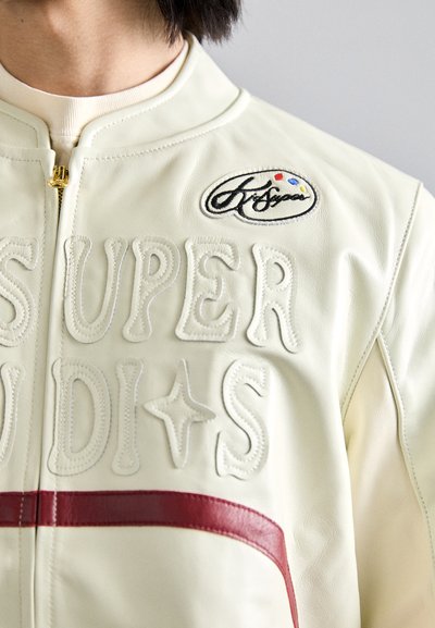 Creme læderjakke med forhøjede tekster "SUPER" og "STUDIOS", en rød stribe og et broderet logo patch. Lynlås lukning foran.