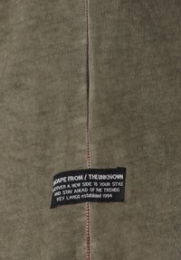 Tejido verde oliva con una textura suave. Presenta costuras rojas a lo largo de la costura y una etiqueta negra con texto blanco que detalla la marca y el lema.