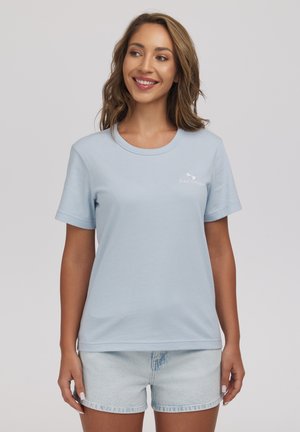 Kobieta w jasnoniebieskiej koszulce z krótkim rękawem i jasnych dżinsowych szortach, uśmiechająca się i patrząca na bok na jednolitym tle.