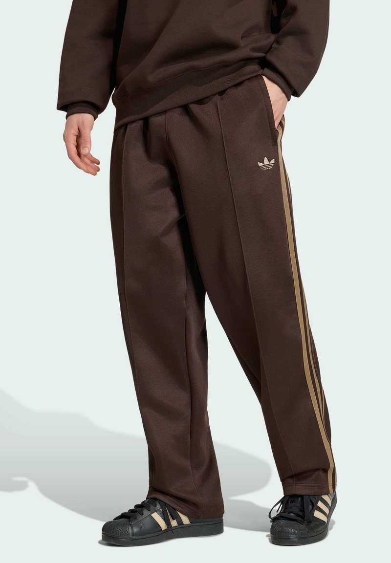 Person, gekleidet in braune Adidas-Trainingshose mit goldenen Streifen und schwarze Adidas-Sneaker mit goldenen Streifen, steht mit einer Hand in der Tasche.