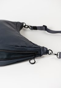 Dunkelgraue, dreieckige Tasche aus weichem, strukturiertem Material. Verfügt über eine Reißverschlusstasche, einen verstellbaren Tragegurt und metallische Akzente.