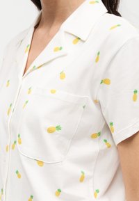 Chemise blanche à manches courtes avec imprimé ananas jaune, poche avant et col ouvert portée par une personne aux cheveux foncés.