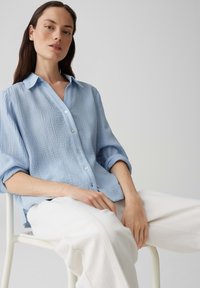 Camicia button-up azzurro chiaro con texture e colletto, con maniche arrotolate, abbinata a pantaloni bianchi, seduto su una sedia.