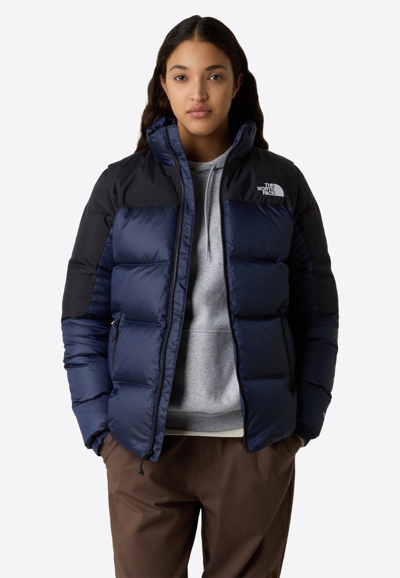 Ung kvinna klädd i en navy- och svart vadderad jacka från The North Face över en grå hoodie och bruna byxor, stående med händerna i fickorna.
