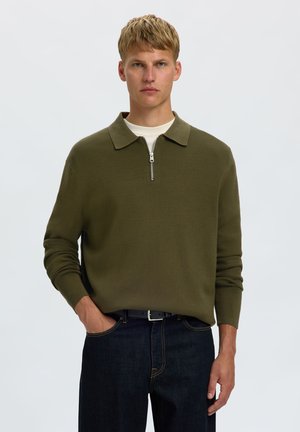 Jeune homme aux cheveux blonds courts porte un pull zippé vert olive sur une chemise blanche et un jean bleu foncé avec une ceinture noire.