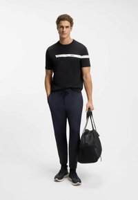 Schwarzes T-Shirt mit weißem Streifen und Logo, kombiniert mit marineblauen Hosen. Das Modell hält eine schwarze Tasche und trägt dunkle Sneakers mit strukturierten Details.