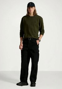 Camisola de manga comprida verde-oliva, calças cargo pretas com bolsos, sapatilhas pretas e um boné preto com um logótipo, em pé sobre um fundo claro.