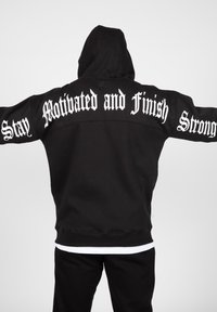 Melns kapucis ar priekšējo kangaroo kabatu, uz aizmugures liela balta teksta: "Paliec motivēts un pabeidz spēcīgi." Klasiska piegriezuma un tekstūras.