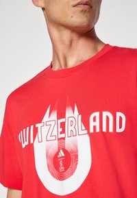 Rød bomulds T-shirt med ordet "SWITZERLAND" i hvid, med et grafisk design der inkluderer konturer og et adidas-logo.