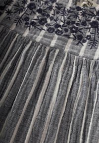 Tissu rayé gris et blanc avec des détails floraux brodés en sombre. La texture est douce avec un design plissé en bas.