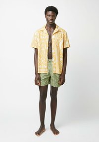 Korte mouwen shirt in geel met een wit patroon, gecombineerd met groene shorts met een subtiel geometrisch print. Informele strandkleding.