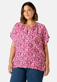 Blommig blus i stark rosa, med korta ärmar och en nyckelhålsutskärning. Tyget har ett mönster med små vita och lila blommor.