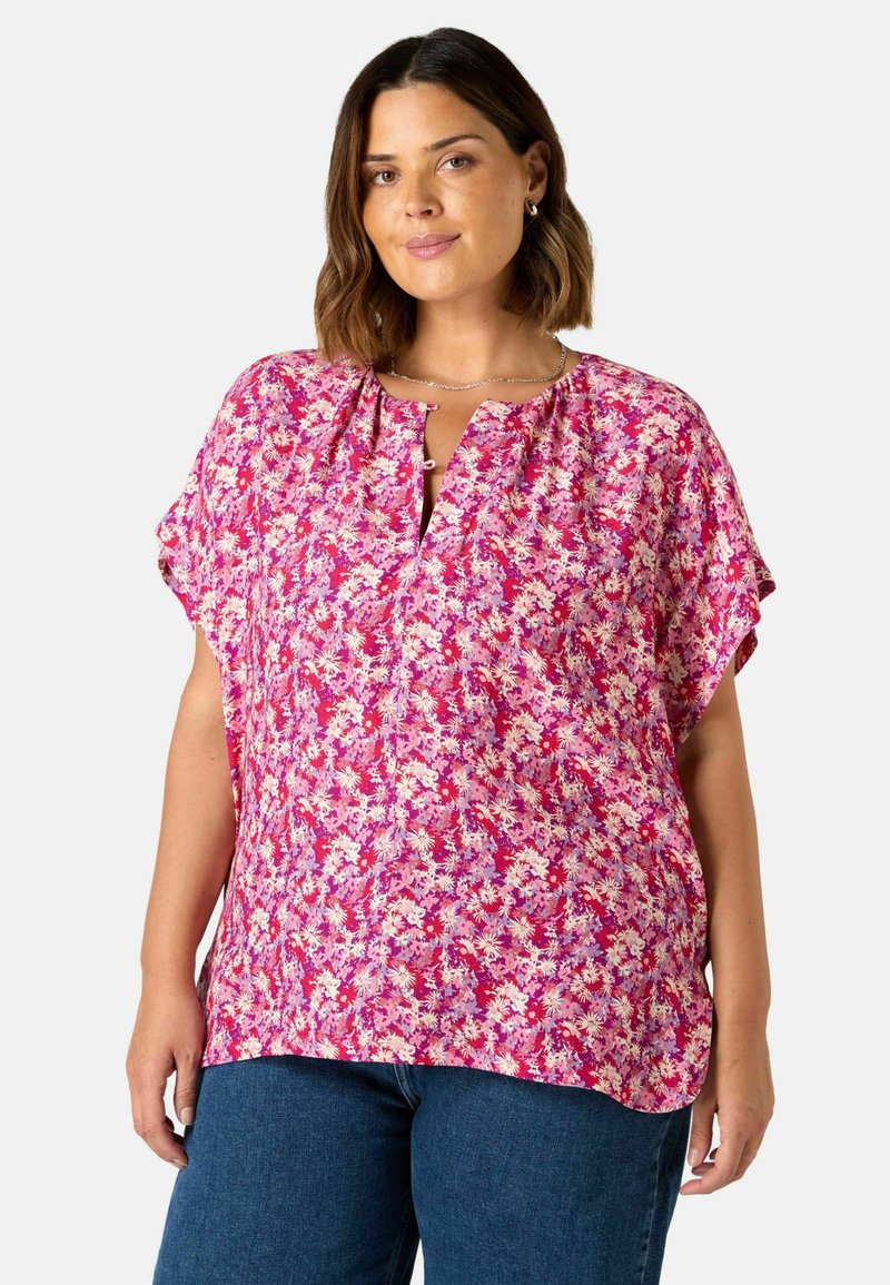 Blommig blus i stark rosa, med korta ärmar och en nyckelhålsutskärning. Tyget har ett mönster med små vita och lila blommor.