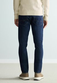 Sötétkék farmernadrág slim fit szabással, hátsó zsebekkel és kontrasztos varrással, bézs lábbelivel párosítva. Textúrált krémszínű pulóver.