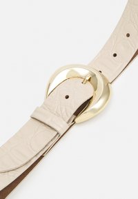 Ceinture en simili cuir beige avec une finition texturée, dotée d'une grande boucle circulaire dorée et de plusieurs trous pour un ajustement réglable.