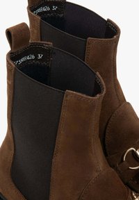 Brązowe zamszowe botki za kostkę z elastycznymi panelami bocznymi, gładką fakturą i metalowymi akcentami. Wewnątrz metka rozmiaru mówi 37.