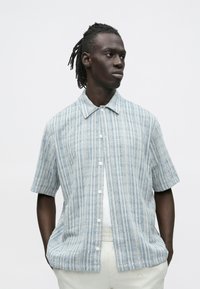 Mees, kellel on dredsid, kannab lühikeste varrukatega helesinist ruudulist nööbitavat särki valge t-särgi ja heledate pükstega, vaatab kõrvale.