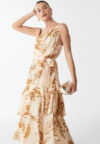 Abito maxi a motivi floreali in crema con motivi a foglia marrone, volants sovrapposti, vita con cintura e una clutch metallica abbinata.