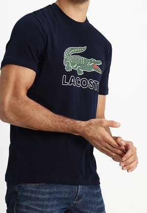 Homme portant un t-shirt bleu marine avec un grand logo crocodile vert et le texte « LACOSTE », assorti à un jean bleu.