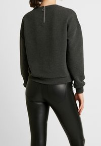 Suéter gris de canalé con cuello redondo y una cremallera corta en la parte de atrás; combinado con pantalones ceñidos de cuero sintético negro.