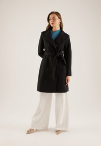 Vrouw draagt een zwart jasje met riem over een blauwe top, witte broek met wijde pijpen en spitse hakken met strikdetails, staand tegen een effen achtergrond.