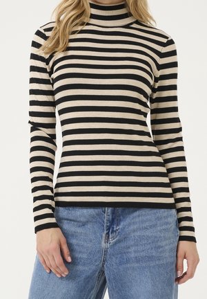 Woman wearing black and beige horizontally striped long-sleeve turtleneck sweater with light blue jeans, standing against a plain background.