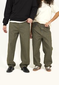 Grüne oversized Jeanshose mit seitlichen Taschen, sichtbaren Nähten und relaxed Fit, kombiniert mit unterschiedlichen Schuhen in einer neutralen Umgebung.