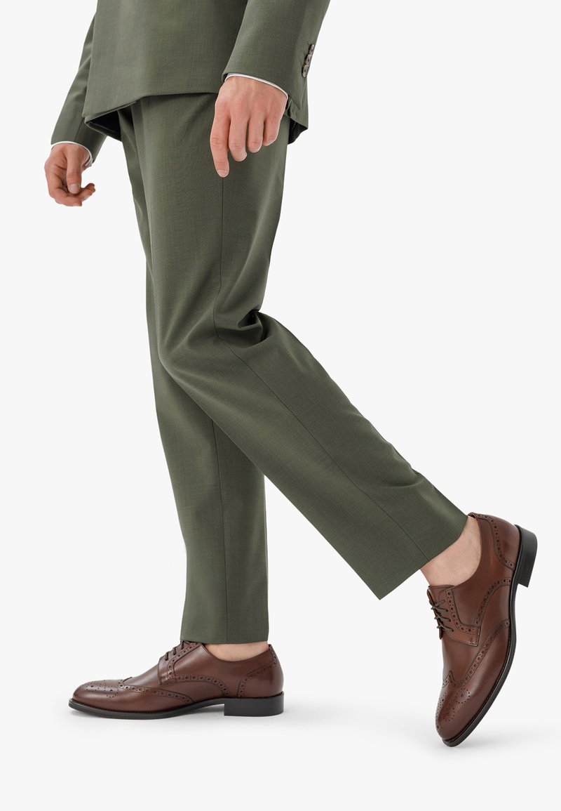 Scarpe eleganti in pelle marrone con dettagli perforati, allacciate, e una superficie liscia, indossate con pantaloni tagliati color verde oliva.