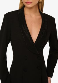 Czarna marynarka dwurzędowa z głębokim dekoltem w serek ozdobiona czarnymi, błyszczącymi detalami. Gładki materiał o wyraźnej strukturze i guziki.