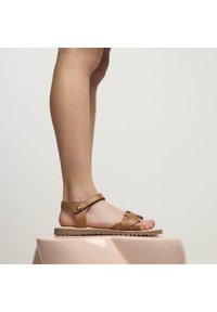 Sandali in pelle color tan con cinturini incrociati e fibbia, dotati di una suola testurizzata. Posizionati su una superficie rosa, che mette in mostra il design e la vestibilità.