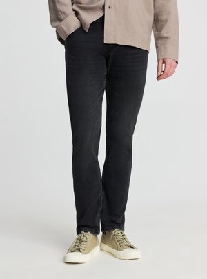 Mężczyzna w czarnych dopasowanych dżinsach, beżowej koszuli zapinanej na guziki i jasnobeżowych trampkach na sznurowadła stoi na jednolitym tle.