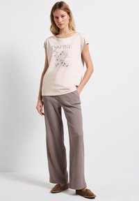 Camiseta de color beige claro con el texto "INSPIRE" y un diseño floral, combinada con pantalones anchos de color marrón con ondas. El modelo lleva zapatos marrones tipo slip-on.