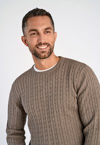 Brun, kabelstickad tröja med rund hals och långa ärmar, texturerat mönster, bärs över en vit skjorta.