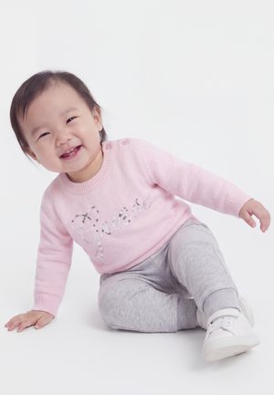 Tout-petit souriant assis par terre portant un pull rose avec le texte "Guess", un pantalon gris et des chaussures blanches sur fond blanc.