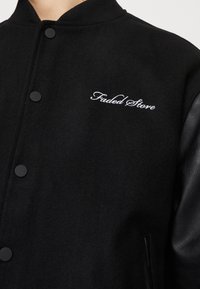 Czarna wełniana kurtka bombowiec z skórzanymi rękawami, czarnymi zatrzaskami i białym haftowanym logo "Faded Store" na klatce piersiowej.