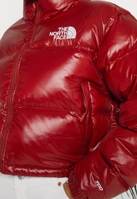 Czerwona kurtka puchowa z błyszczącym wykończeniem, zamek błyskawiczny, logo na klatce piersiowej, kieszeń boczna i elastyczne mankiety. Posiada izolację o mocy 700 fill power.