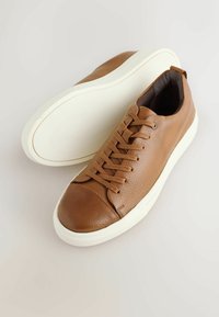 Zapatillas de cuero marrón con un acabado texturizado, que presentan una puntera redondeada, un diseño de cordones y una suela de goma blanca en contraste.